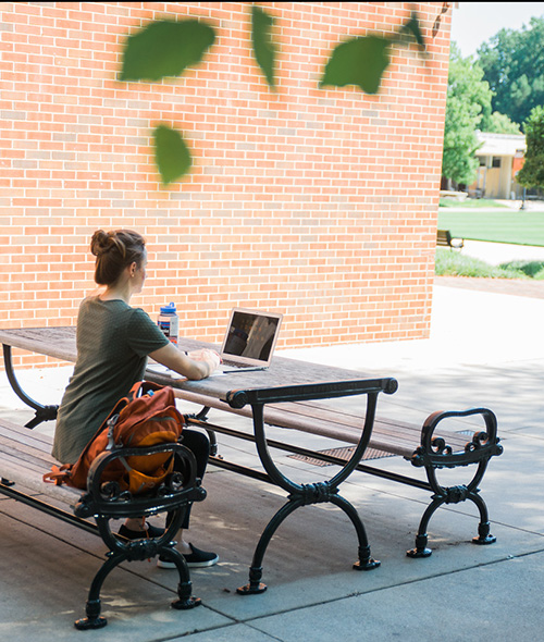 student sitting at a bench and table outside and working on a laptop